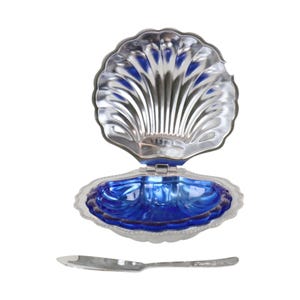 May include: A silver-toned metal clam shell dish with a hinged lid that opens to reveal a deep blue glass insert. The shell features a scalloped edge and a ribbed interior design. A matching silver-toned metal spreader knife is placed below the dish.