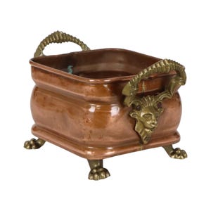 May include: A square copper container with a polished finish, featuring two brass handles shaped like ram heads with curved horns. The vessel is supported by four brass feet designed to resemble animal paws, providing a stable base.