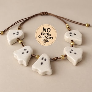 May include: A bracelet featuring five off-white ceramic ghost charms with black painted eyes and mouths, strung on a brown cord with small gold-toned metallic beads. A circular sticker in the centre displays the text NO EXTRA CUSTOMS FEES.