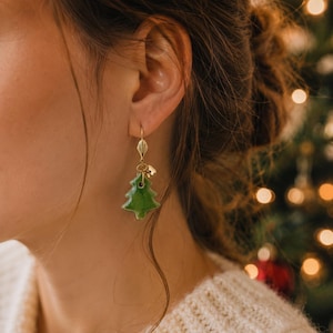 Op de afbeelding: Een goudkleurige oorbel met een kleine, glanzend groene keramische kerstboomhanger die aan een delicaat metalen haakje hangt. De hanger is voorzien van een piepklein goudkleurig belletje dat aan de verbindingsring is bevestigd.