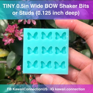 以下が含まれることがあります： ターコイズブルーのシリコンモールドで、9つのリボン型のキャビティがあります。各リボンは幅1.27cm、深さ0.32cmです。モールドは、ピンクの花が咲く緑色の背景に、手で持たれています。