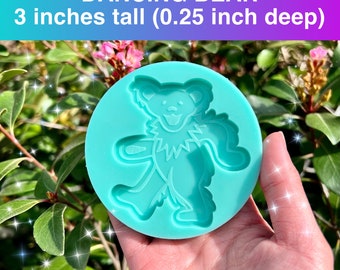 Puede incluir: Un molde de silicona de color turquesa con un diseño de oso bailarín. El molde redondo mide 7,6 cm de alto y 0,6 cm de profundidad. El texto "DANCING BEAR" se muestra en la parte superior. El molde se sostiene en una mano sobre un fondo de follaje verde.