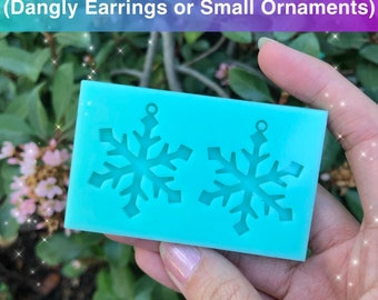 May include: A rectangular turquoise silicone mold featuring two snowflake-shaped cavities with small circular loops at the top for hanging. The text 1.5 inches Snowflakes Mold (Dangly Earrings or Small Ornaments) is printed at the top.