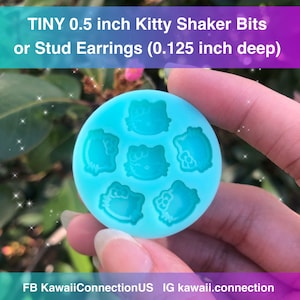 Op de afbeelding: Een lichtblauwe, cirkelvormige mal met zes kleine Hello Kitty-ontwerpen. De tekst op de afbeelding luidt "TINY 0.5 inch Kitty Shaker Bits or Stud Earrings (0.125 inch deep)". De mal wordt in een hand gehouden. De diameter is 1,27 cm.