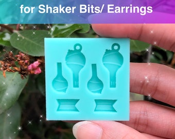 May include: A light blue silicone mold with four key-shaped cavities and two rectangular cavities. The mold is designed to create shaker bits or earrings with a BTS logo and light bomb design. The text "TINY BTS Logo & Light Bomb for Shaker Bits/Earrings" is at the top of the image. The text "FB KawaiiConnectionUS IG kawaii.connection" is at the bottom of the image.