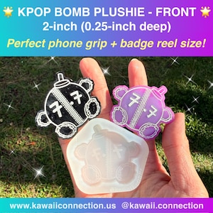 May include: A KPOP Bomb Plushie in black and purple, with the text "7.7" on the front. The plushie is 2 inches in size and is designed as a phone grip or badge reel. The image also shows a clear mold of the plushie.