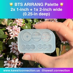 以下が含まれることがあります： 手で持たれた、透明な長方形のクラフト用モールド。 モールドには「BTS ARIRANG Palette」とラベルが付けられ、円形と長方形の形状が含まれています。 サイズは2x 2.54cm、幅1x 5.08cm、深さ0.64cmです。 画像にはウェブサイトとソーシャルメディアハンドルも表示されています。