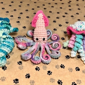 Op de afbeelding: Drie gehaakte zeedieren: een blauw en groen zeepaardje, een roze en paarse octopus en een roze en groene kwal.