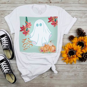 May include: White t-shirt featuring a graphic of a ghost, pumpkins, bats, and flowers on a light blue background.