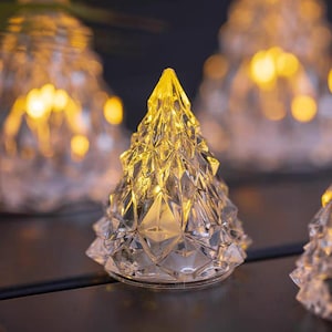 Puede incluir: Luces decorativas con forma de árbol de Navidad de cristal transparente. Los árboles tienen un diseño facetado y están iluminados desde dentro, creando un brillo cálido y dorado. Las luces están sobre una superficie oscura, con otras luces similares en el fondo.