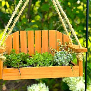 A small wooden planter shaped like a miniature park bench, hung by thick jute rope outside with green leafy plants growing inside against a blurred green foliage background.
