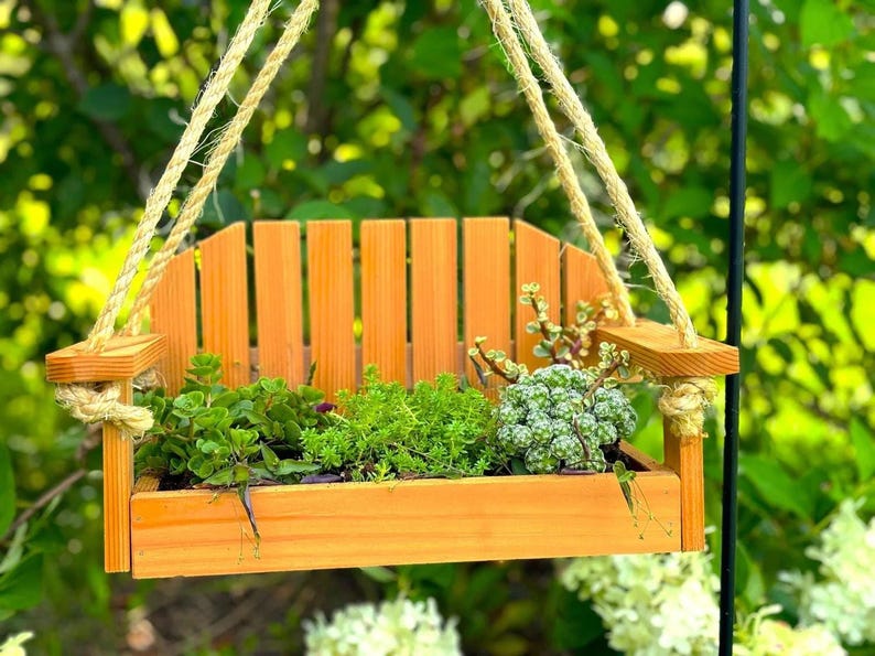 A small wooden planter shaped like a miniature park bench, hung by thick jute rope outside with green leafy plants growing inside against a blurred green foliage background.