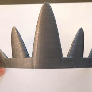A close-up front view of a gray 3D-printed crown with five conical spikes of varying heights, held by a hand on the left against a white background.