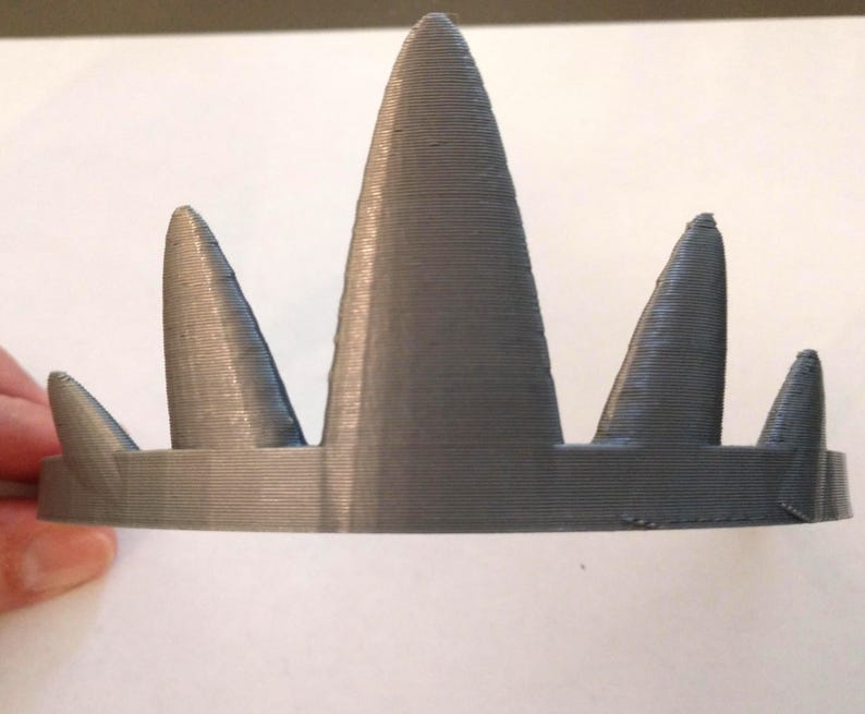 A close-up front view of a gray 3D-printed crown with five conical spikes of varying heights, held by a hand on the left against a white background.