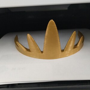 A gold 3D-printed crown resting on a white surface inside a dark enclosure, displaying its horizontal print lines and pointed spikes.