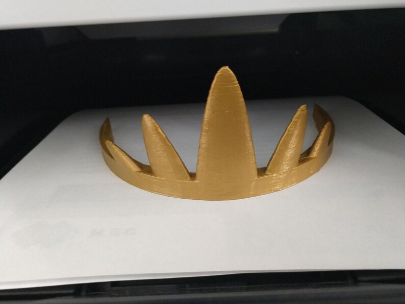 A gold 3D-printed crown resting on a white surface inside a dark enclosure, displaying its horizontal print lines and pointed spikes.