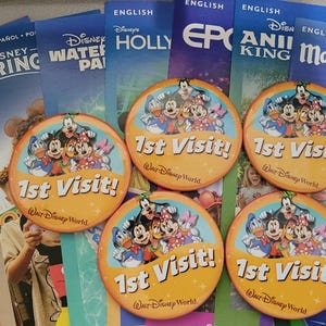 Może przedstawiać: Cztery okrągłe żółte przypinki Walt Disney World 1st Visit z Myszką Miki, Myszką Minnie, Kaczorem Donaldem i Goofym, ułożone na kolekcji kolorowych map i broszur parków rozrywki Disney, w tym Magic Kingdom i ESPN Wide World.