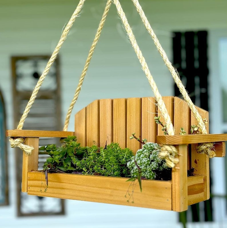 A wooden swing-style planter box holding lush green succulents, suspended by thick rope against a neutral blurred background.