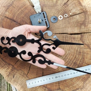 May include: Black clock hands with a decorative, ornate design. The hands are made of metal and have a black finish. The hands are shown on a wooden surface with a ruler for scale.