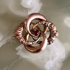 May include: A gold-coloured brooch with an intricate design. The brooch features three interlocking circular elements, with ornate detailing on some sections. A small, round, red gemstone is set in the centre. The pin is a vintage-style accessory.