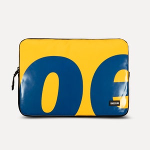 May include: Yellow laptop sleeve with a blue "oe" design and the word "UNBEGUN" in black text.