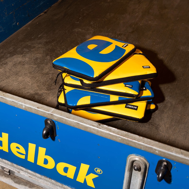 May include: A stack of four yellow and blue padded laptop cases with the word "L&Aring;NGEDR&Auml;KT" printed on one of the cases.