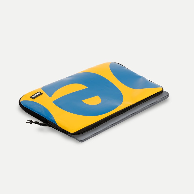 May include: Yellow laptop sleeve with a blue "@" symbol. The sleeve has a black zipper and a black pull tab. The sleeve is lying on a gray surface with a laptop inside.