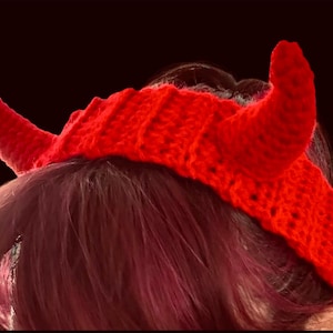 May include: A bright red crocheted headband featuring two prominent, stuffed, cone-shaped horns extending upward from the top. The headband is constructed with a textured, ribbed stitch pattern and sits horizontally across the top of the head.