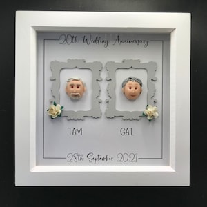 Könnte beinhalten: Ein weißer Rahmen mit grauem Hintergrund und zwei grauen Bilderrahmen im Inneren. Die Bilderrahmen haben weiße Blumen unten und eine Tonbüste eines menschlichen Gesichts im Inneren. Der Text "20th Wedding Anniversary" befindet sich oben am Rahmen. Der Text "TAM" und "GAIL" befindet sich unter den Bilderrahmen. Das Datum "28th September 2021" befindet sich unten am Rahmen.