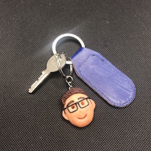 May include: A blue leather keyring with a silver key ring and a small, brown, clay figurine of a person wearing glasses.