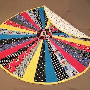 May include: A round, multi-colored quilt with a Mickey Mouse design in the center. The quilt is made of various fabrics with Mickey Mouse patterns, stripes, and polka dots. The quilt is edged with a yellow border.