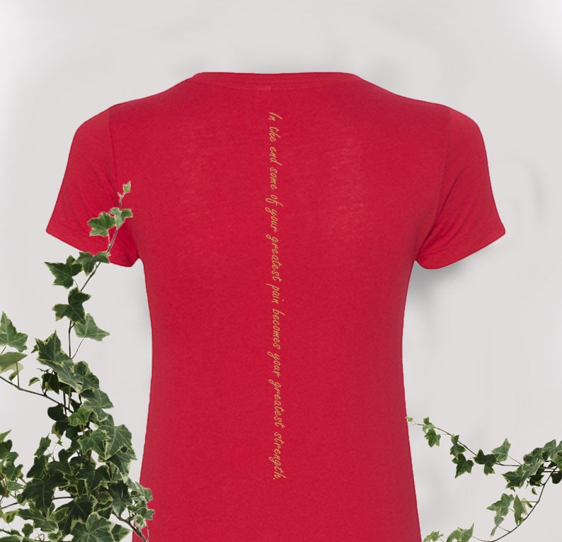 May include: Red short-sleeved t-shirt with a vertical quote in gold lettering on the back: "In the end, some of your greatest pain becomes your greatest strength."