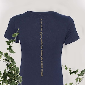 May include: A navy blue t-shirt with short sleeves and a crew neck. The back of the shirt has the text "In the end some of your greatest pain becomes your greatest strength" printed vertically in gold.
