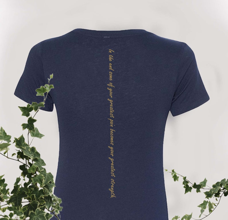 May include: A navy blue t-shirt with short sleeves and a crew neck. The back of the shirt has the text "In the end some of your greatest pain becomes your greatest strength" printed vertically in gold.