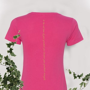 May include: Pink t-shirt with a short sleeve and a yellow quote on the back that reads "In the end some of your greatest pain becomes your greatest strength."