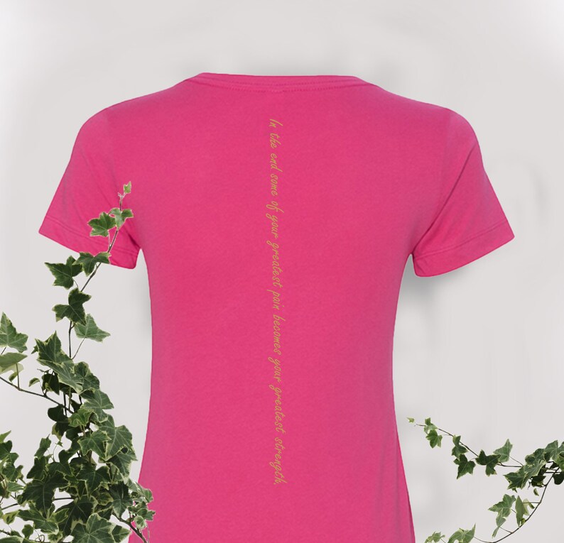 May include: Pink t-shirt with a short sleeve and a yellow quote on the back that reads "In the end some of your greatest pain becomes your greatest strength."