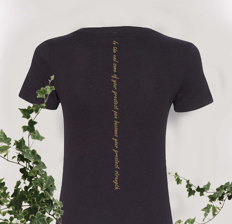 May include: Black t-shirt with a gold inscription that reads "In the end, some of your greatest pain becomes your greatest strength."