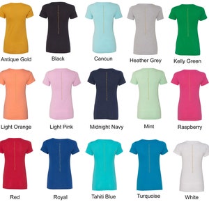 May include: A collection of 15 women's t-shirts in various colors, including antique gold, black, light orange, light pink, midnight navy, mint, raspberry, red, royal, tahiti blue, turquoise, and white. Each shirt features a gold vertical line down the center of the back.