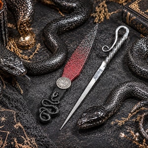 May include: A dark, gothic-themed still life featuring black snake sculptures, a candle, and a decorative dagger with a red and black blade. A silver letter opener and a book with gold accents are also present, creating a mystical aesthetic.