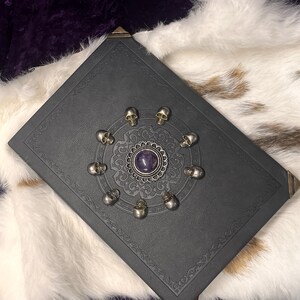May include: A dark gray book with a decorative silver skull and amethyst design. The book has ornate corner accents and is set against a white and brown fur-like background.