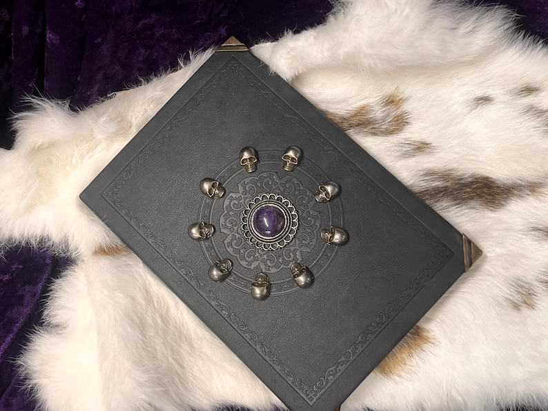 May include: A dark gray book with a decorative silver skull and amethyst design. The book has ornate corner accents and is set against a white and brown fur-like background.