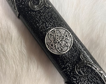 May include: Close-up of a black leather scabbard with silver-toned metal accents. The scabbard features an intricate design with a circular emblem of three overlapping pentagrams.