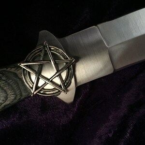 May include: A silver pentagon with a circle around it, on a black and grey wooden handle of a knife. The knife has a silver blade and the text "PAKISTAN" is visible on the blade.