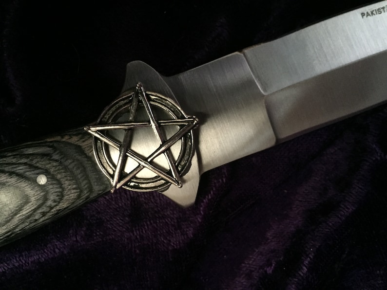 May include: A silver pentagon with a circle around it, on a black and grey wooden handle of a knife. The knife has a silver blade and the text "PAKISTAN" is visible on the blade.