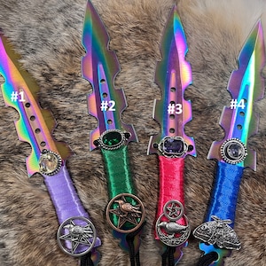 May include: Four iridescent daggers with decorative handles. Each dagger has a different colored handle and a different gemstone at the top. The handles are decorated with a variety of charms, including a crow, a pentagram, and a moth.