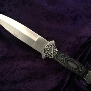 May include: A silver dagger with a black wood handle and a pentagram design on the pommel. The dagger is lying on a purple velvet surface.