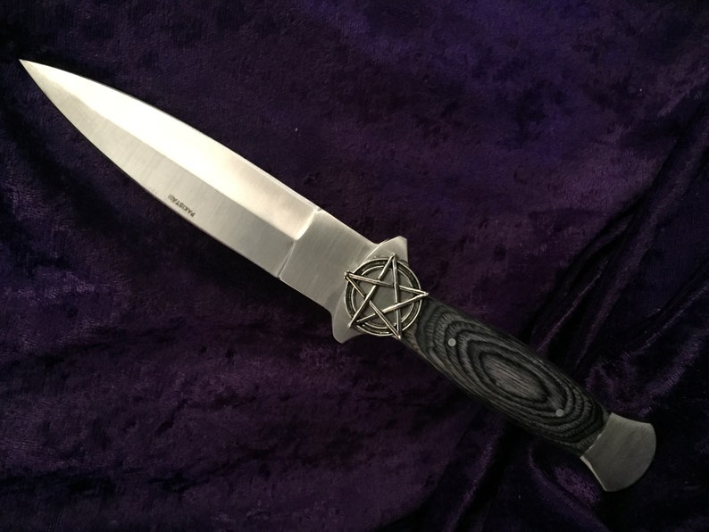 May include: A silver dagger with a black wood handle and a pentagram design on the pommel. The dagger is lying on a purple velvet surface.
