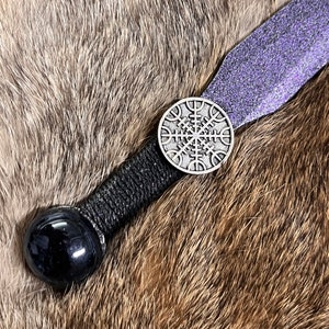 May include: A black and silver leather-wrapped paddle with a purple glitter finish. The paddle has a black obsidian-like sphere at one end and a silver metal circular charm with a Norse Vegvisir symbol at the other end.