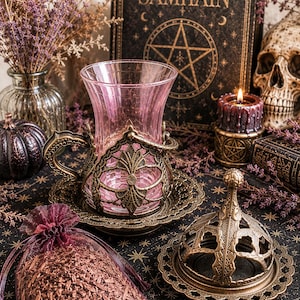 Puede incluir: Una taza de cristal rosa dentro de un soporte de metal color bronce con su plato a juego, acompañada de una tapa de metal con un patrón decorativo recortado y una pequeña bolsa de organza llena de material botánico seco.