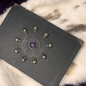 May include: A black leather-bound book with decorative corner protectors. The cover features a central silver medallion with a purple stone, surrounded by a circle of small silver skulls. The book rests on a white and brown fur-like surface.